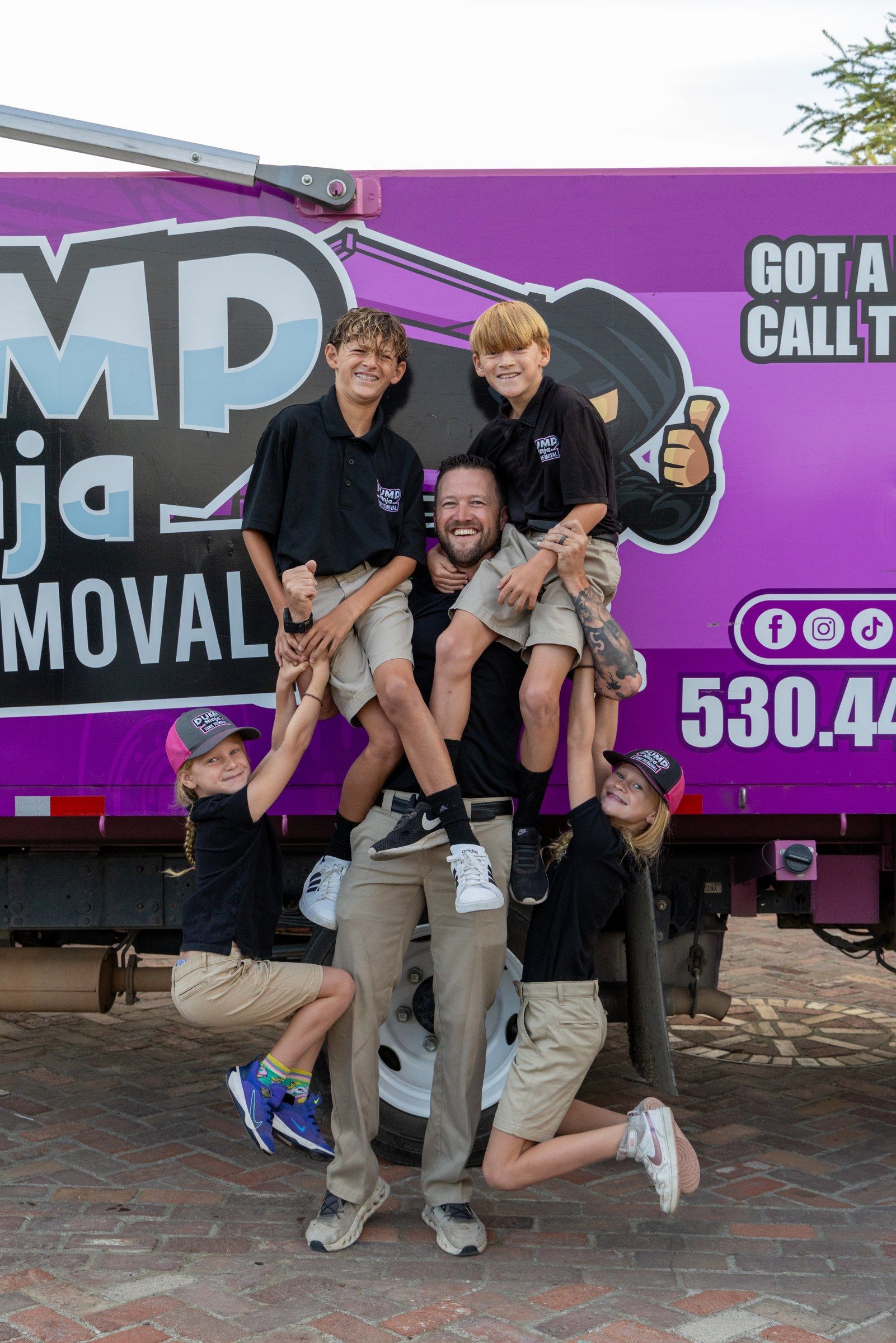 Jeremy with his four children climbing on his shoulders and holding his arms beside the Dump Ninja truck