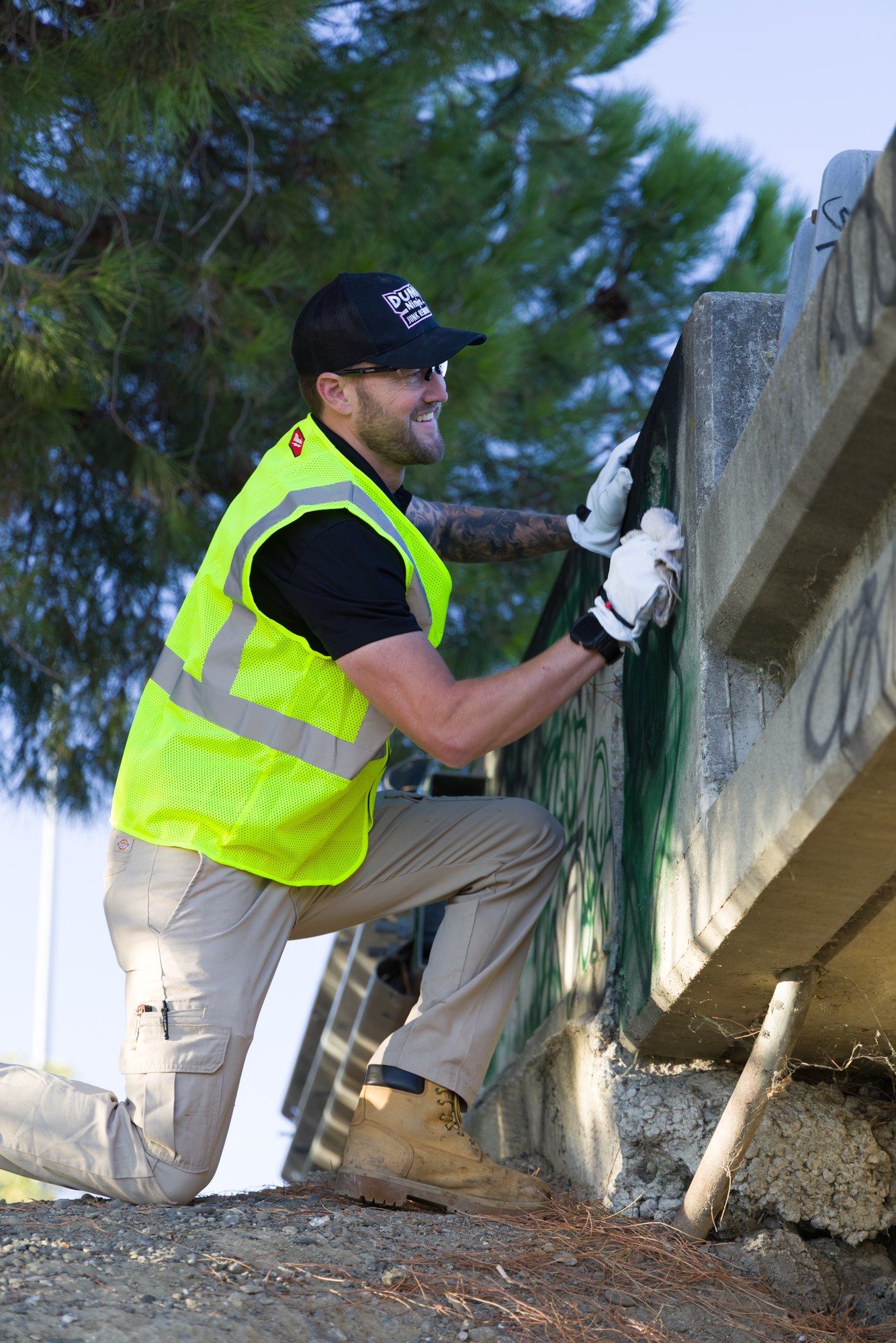 A Dump Ninja crew member in a high-visibility vest removing graffiti from a concrete overpass wall