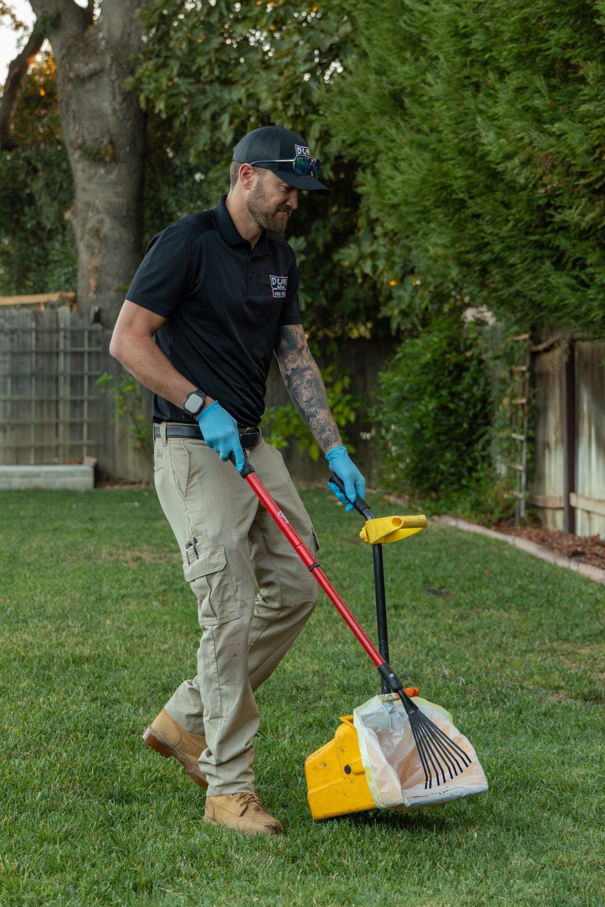 Dump Ninja crew member scooping dog waste along a fence line with a rake and pan in a Woodland backyard